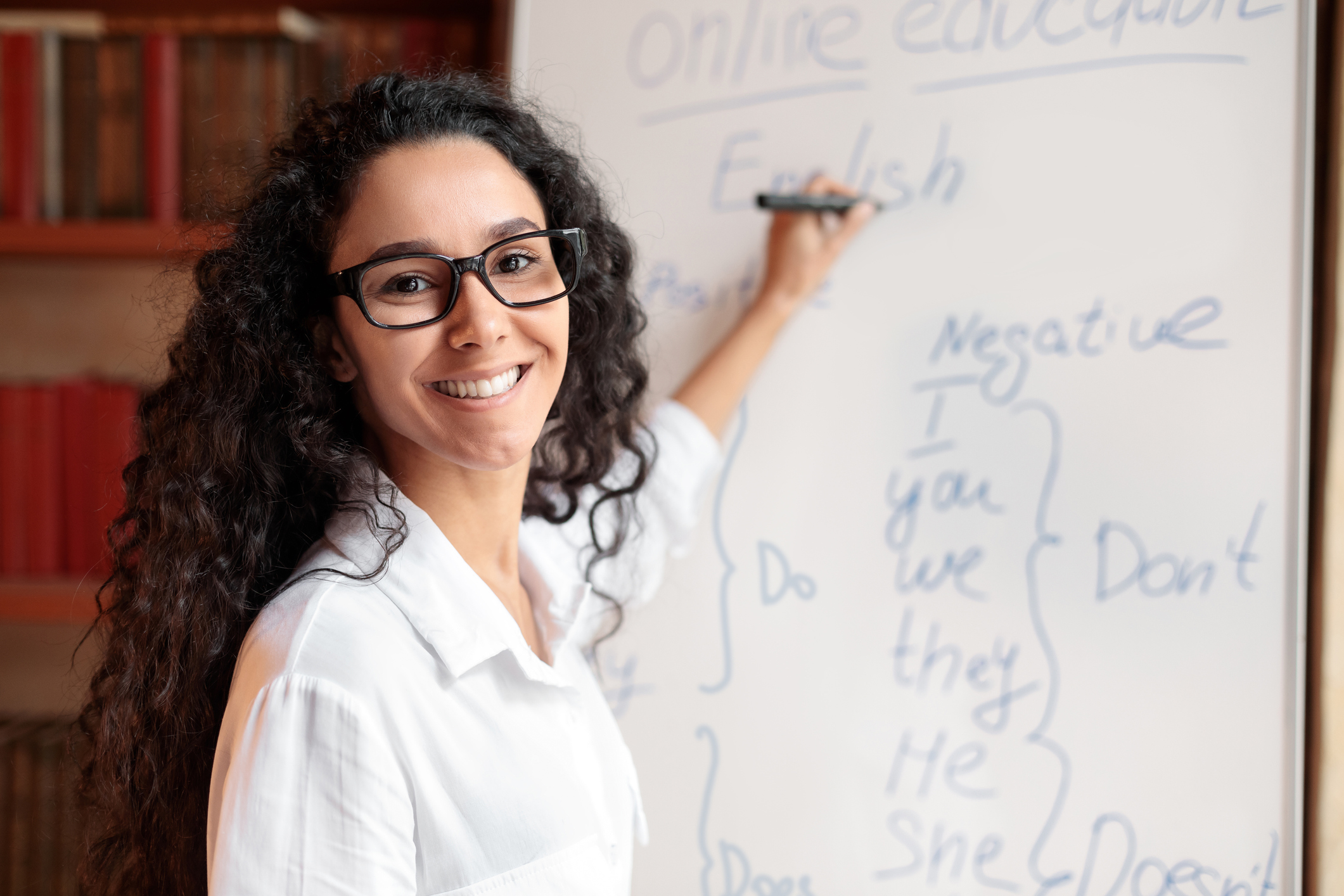 Education And Learning Concept. Portrait of smiling female teacher standing at whiteboard, explaining grammar rules to students. Excited woman in glasses looking at camera, writing on the board